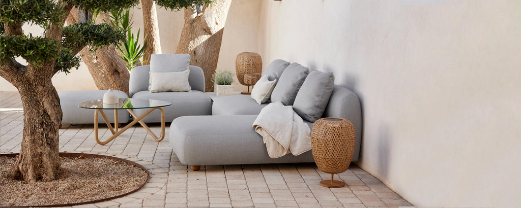 Área de estar elegante con suaves sofás grises, cojines decorativos, una manta acogedora y elementos naturales como un árbol en maceta y mesas de acento tejidas.