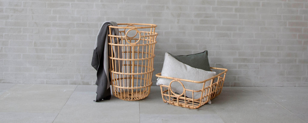 Two woven baskets in natural tones, one large and tall with a grey blanket draped over it, and a smaller one holding soft cushions.