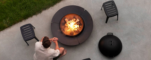 A person sits near a circular fire pit surrounded by modern black chairs, with flames and embers glowing in the center.