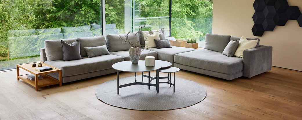 Una sala de estar espaciosa y moderna con un gran sofá seccional gris, una alfombra redonda y mesas de café elegantes, con vegetación visible afuera.