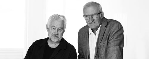 Dos hombres posan juntos con expresiones amigables, vestidos con ropa casual contra un fondo claro, transmitiendo colaboración y creatividad.