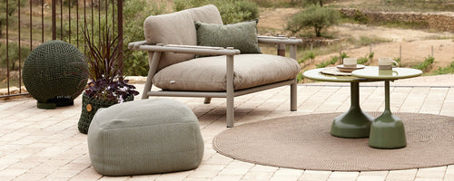 Área de estar elegante con una silla cómoda, mesa redonda, pouf y accesorios decorativos dispuestos sobre una alfombra texturizada.