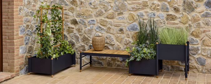 Una exhibición de macetas elegantes con diversas plantas verdes, complementada por un banco de madera contra una pared de piedra rústica.