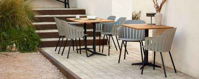 Un ambiente de café elegante con mesas de madera y sillas grises dispuestas en un área pavimentada, complementado por vegetación en el fondo.