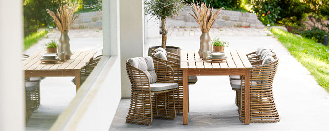 una mesa de madera elegante rodeada de sillas tejidas, con plantas decorativas y vegetación al fondo, perfecta para reuniones.