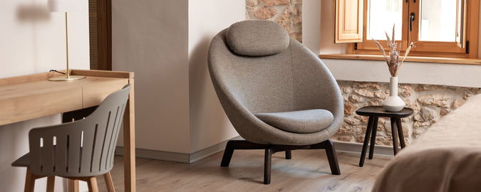 Área de descanso acogedora con una silla moderna de diseño redondeado, una pequeña mesa auxiliar y un escritorio minimalista. La luz natural entra por una ventana.