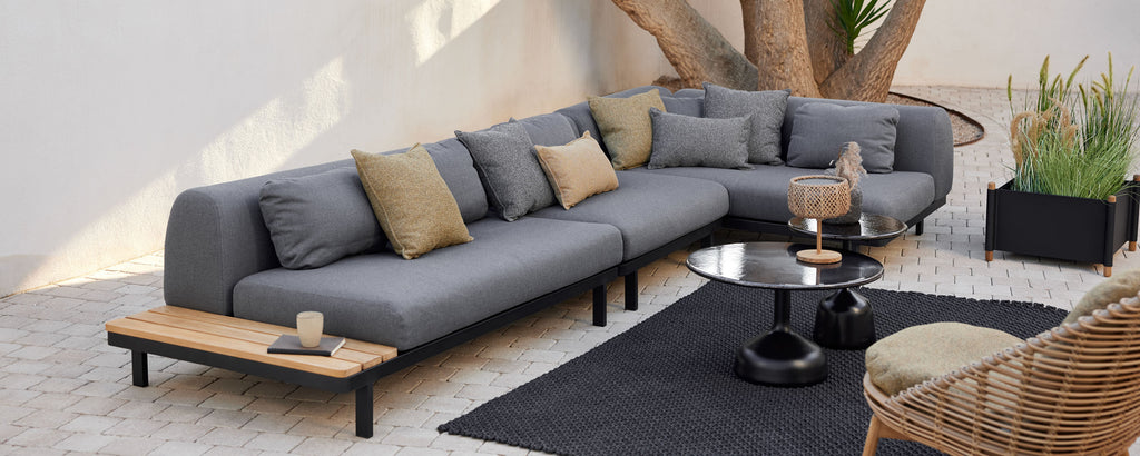 Área de lounge con un sofá seccional moderno con cojines decorativos, una mesa de café redonda y una alfombra elegante, complementada con vegetación.