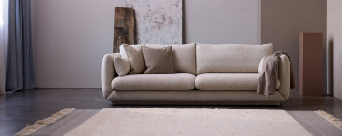 Sofá beige moderno con cojines y una manta gris en un entorno de salón minimalista.