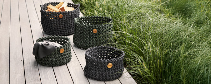 Colección de cestas de mimbre negras dispuestas en una terraza de madera con vegetación de fondo.
