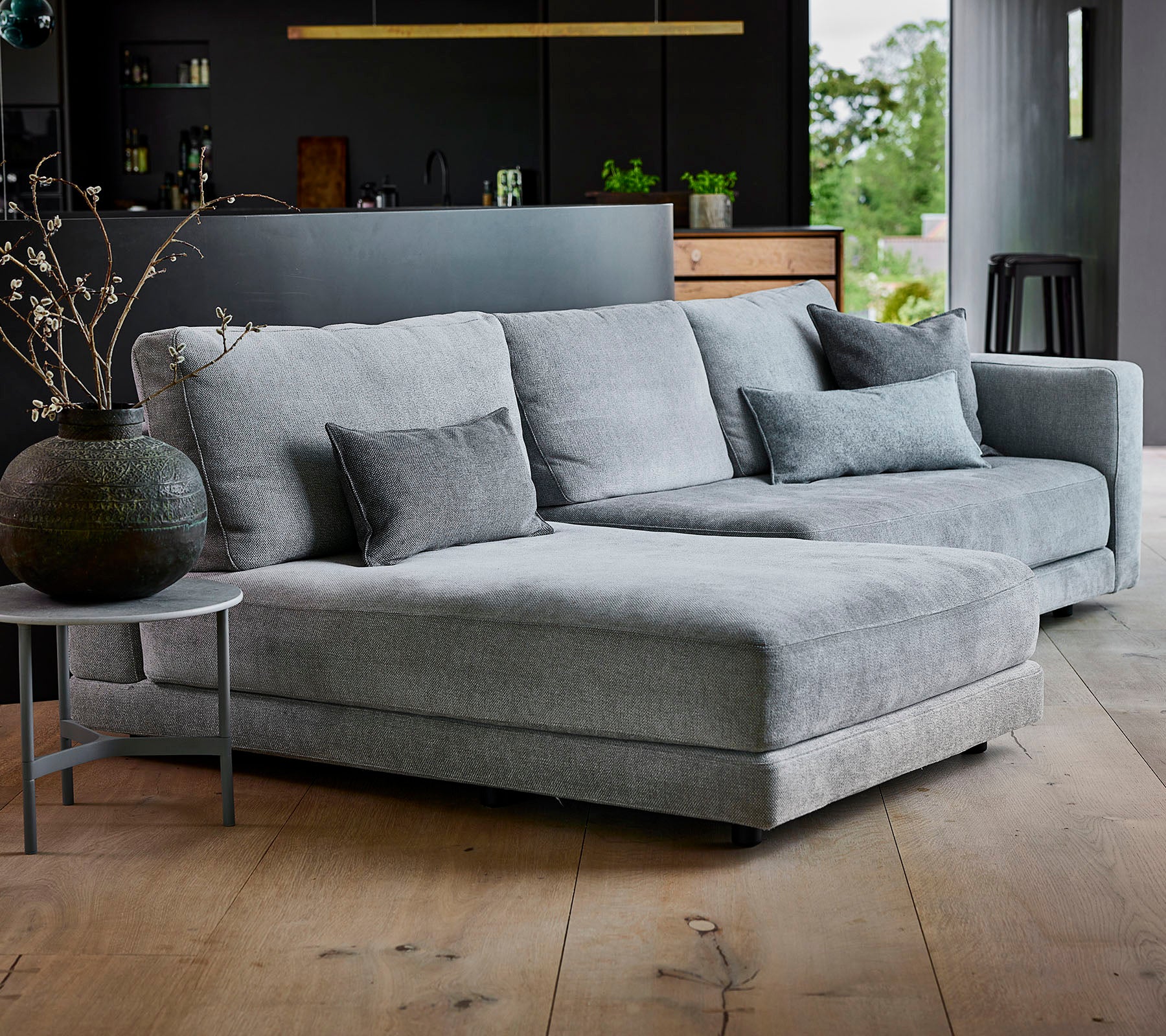 Sofá seccional gris con tela texturizada y cojines decorativos, acompañado de una mesa auxiliar redonda y un jarrón elegante.