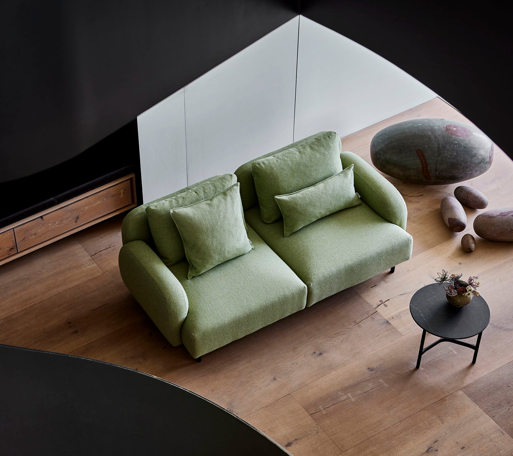 Sofá verde elegante con cojines mullidos al lado de una pequeña mesa redonda, situado contra un fondo moderno con suelo de madera y piedras lisas.