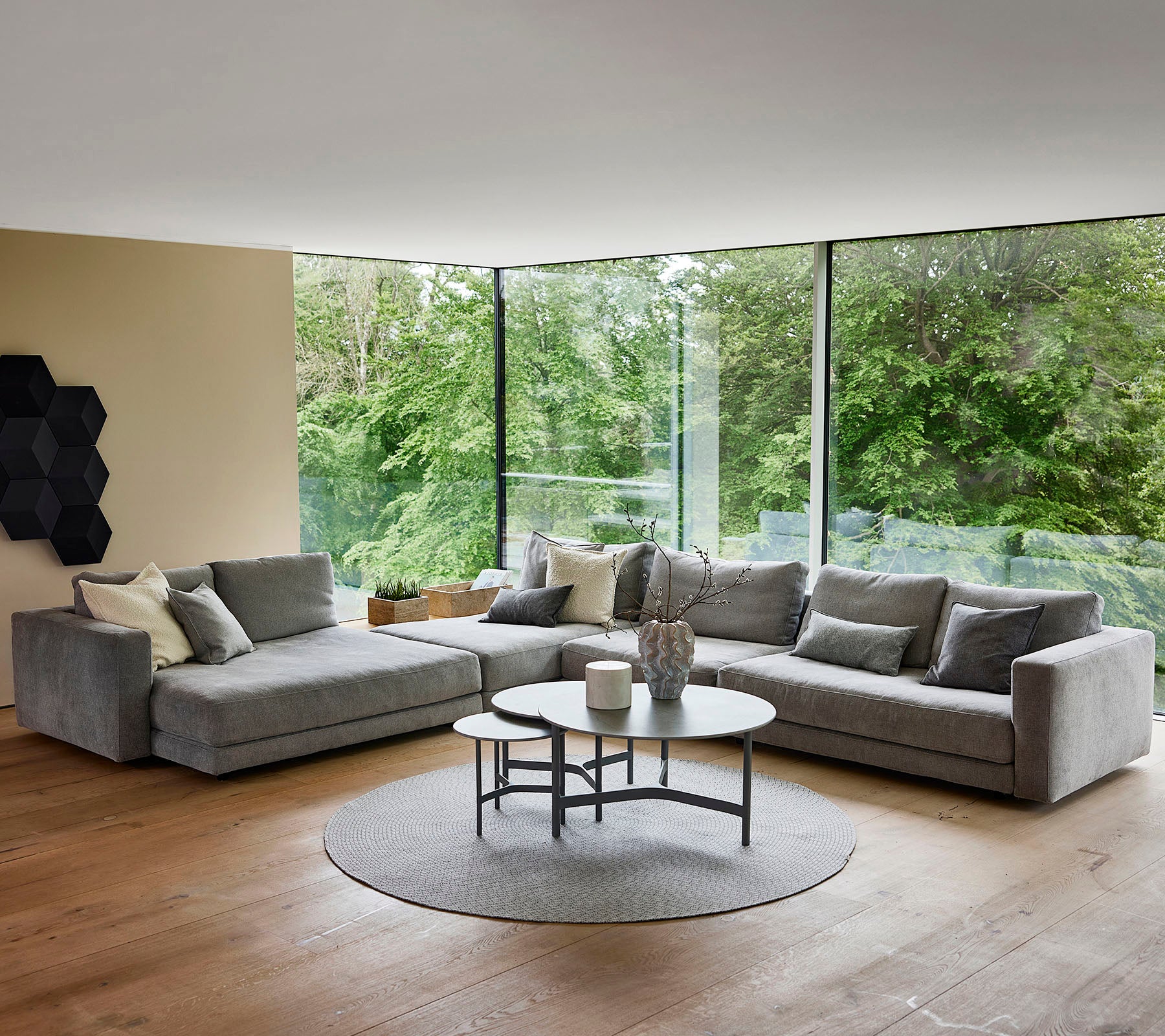 Una sala de estar moderna con un sofá seccional gris con cojines decorativos, una mesa de café redonda y una alfombra circular, rodeada de vegetación.