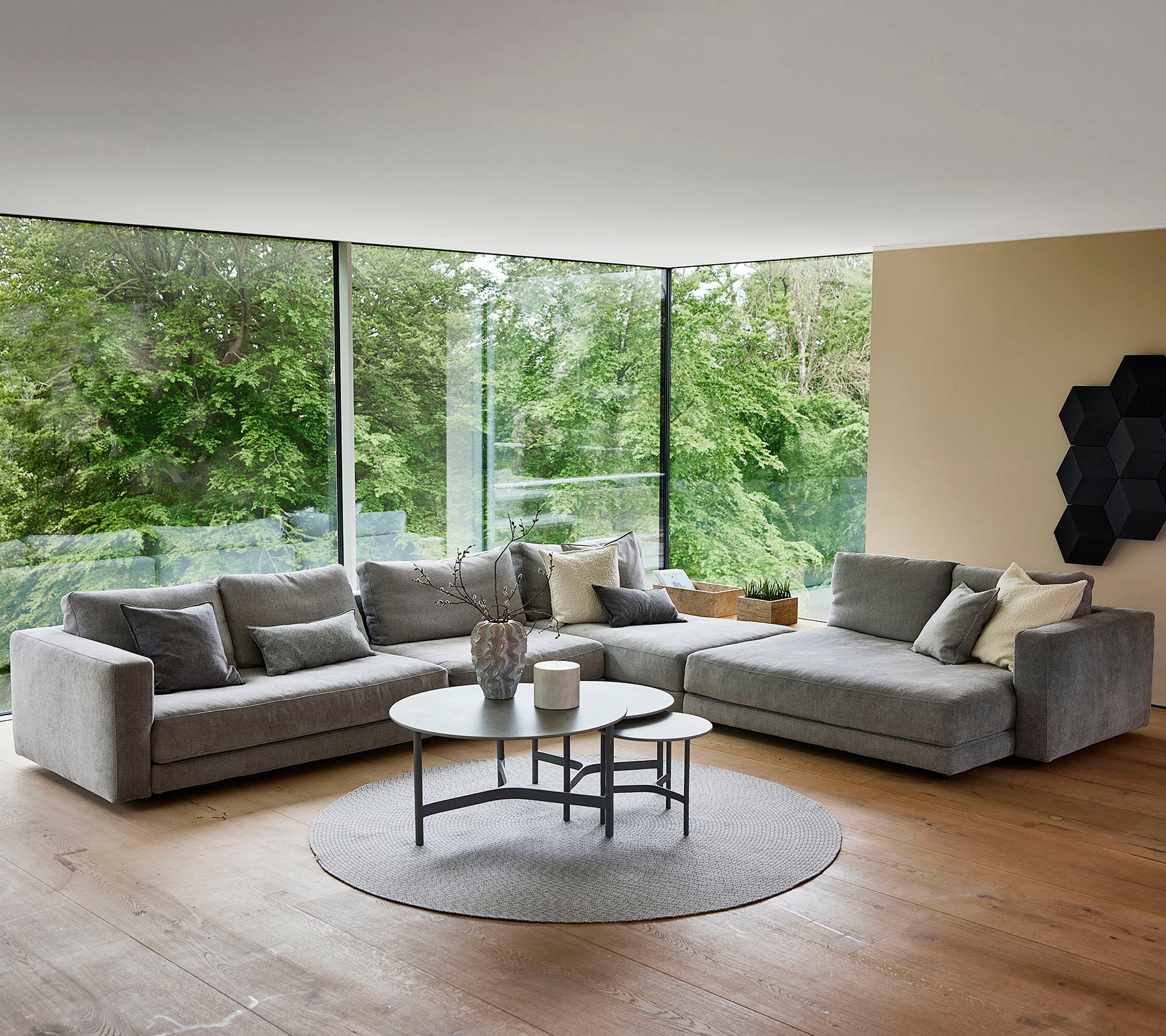 Un espacio de vida moderno y elegante que cuenta con un sofá seccional gris de felpa, una mesa de café redonda y grandes ventanales que muestran la vegetación exterior.