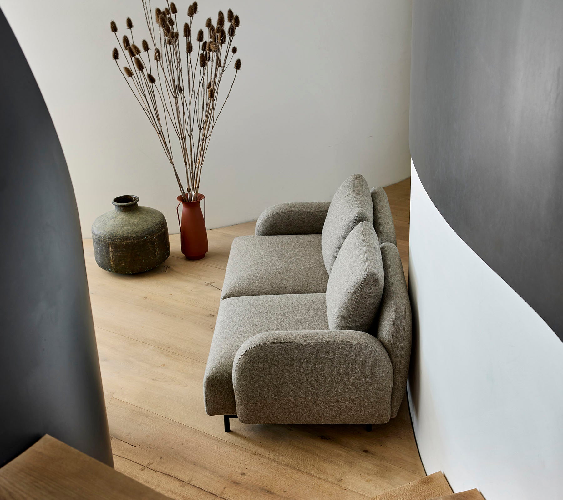 Un sofá beige acogedor situado al lado de un jarrón decorativo y plantas secas, en una pared curva y un suelo de madera.