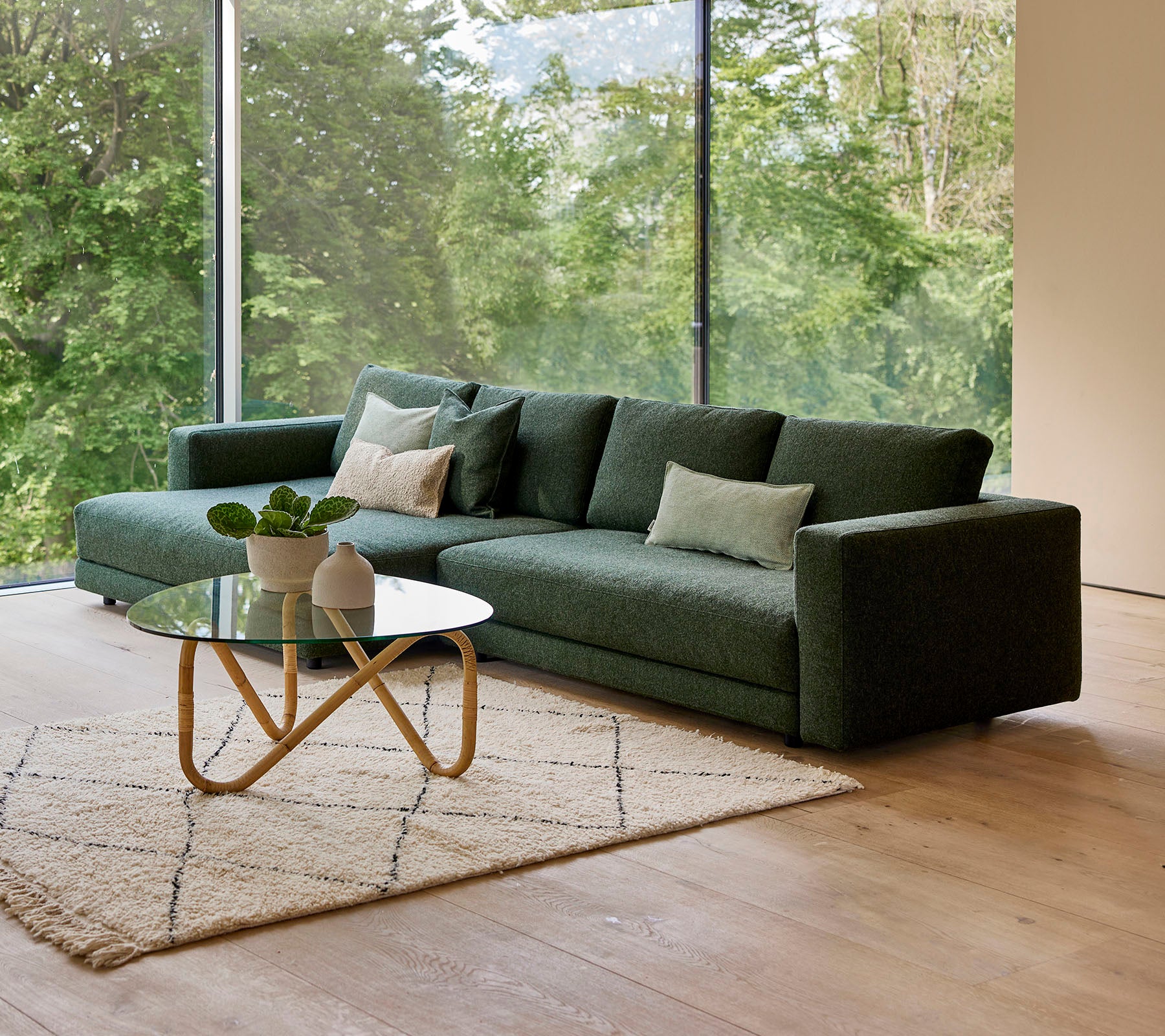 Un sofá verde moderno con almohadas, acompañado de una mesa de café redonda y una alfombra decorativa, con un fondo de vegetación.