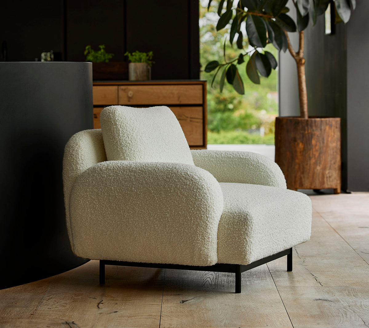 Silla de lounge blanca y acogedora con reposabrazos redondeados y una textura suave, situada frente a un fondo de vegetación y muebles modernos.