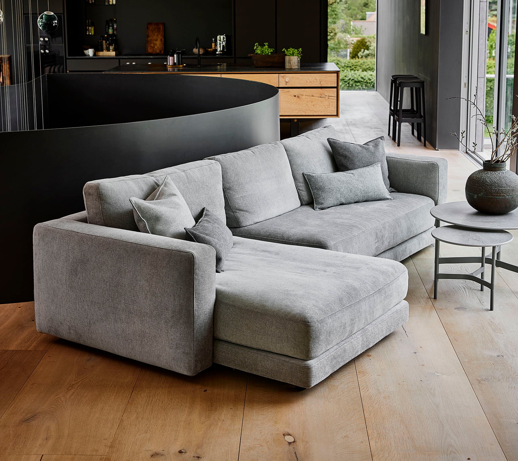 Sofá seccional gris moderno con múltiples cojines, acompañado de una mesa de café redonda y un jarrón decorativo. Diseño simple y elegante.