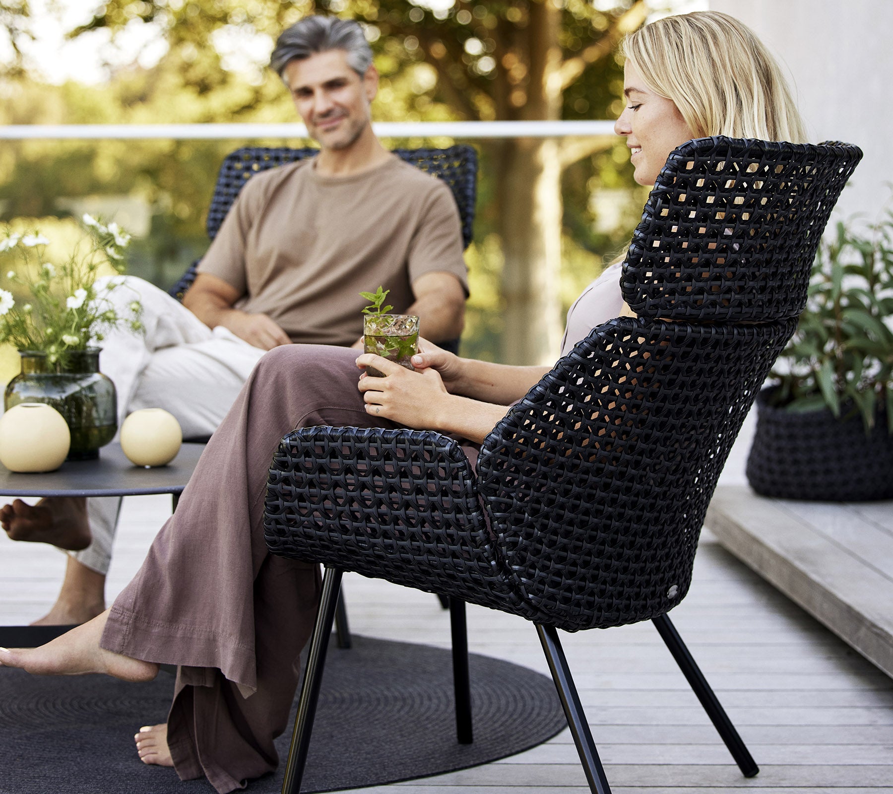 Una silla alta y elegante de material tejido oscuro, con dos personas disfrutando de un momento relajado rodeados de plantas y decoración.