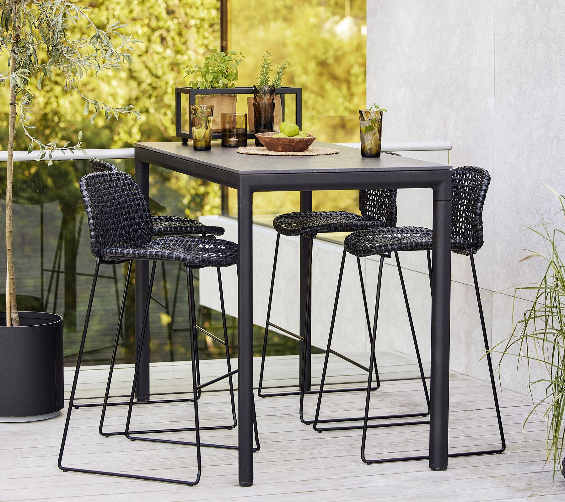 Un elegante conjunto de mesa de bar con tres taburetes de mimbre, dispuesto con vajilla y bebidas, rodeado de vegetación.