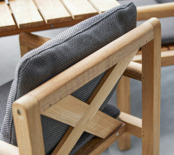 Silla de madera con un cojín de patrón gris, que presenta un diseño en cruz en el respaldo y rodeada de listones de madera.