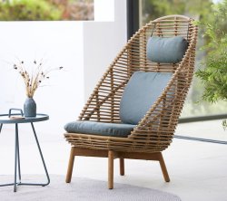 Silla de mimbre elegante con respaldo alto, asiento acolchado y cojín a juego, acompañada de una mesa pequeña y un jarrón decorativo.