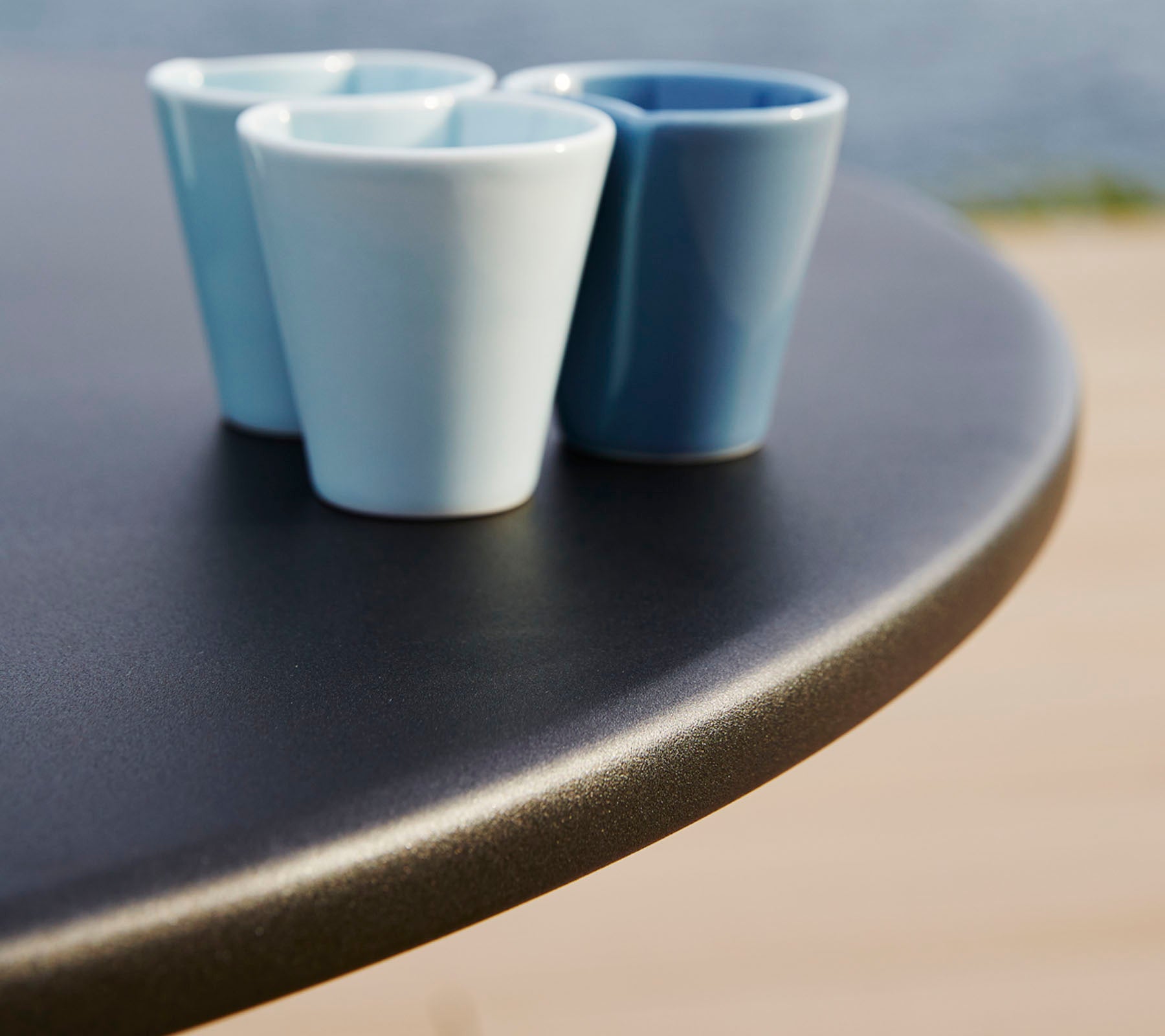 Tres pequeñas tazas de cerámica en diferentes tonos de azul están sobre una superficie de mesa redonda y suave.