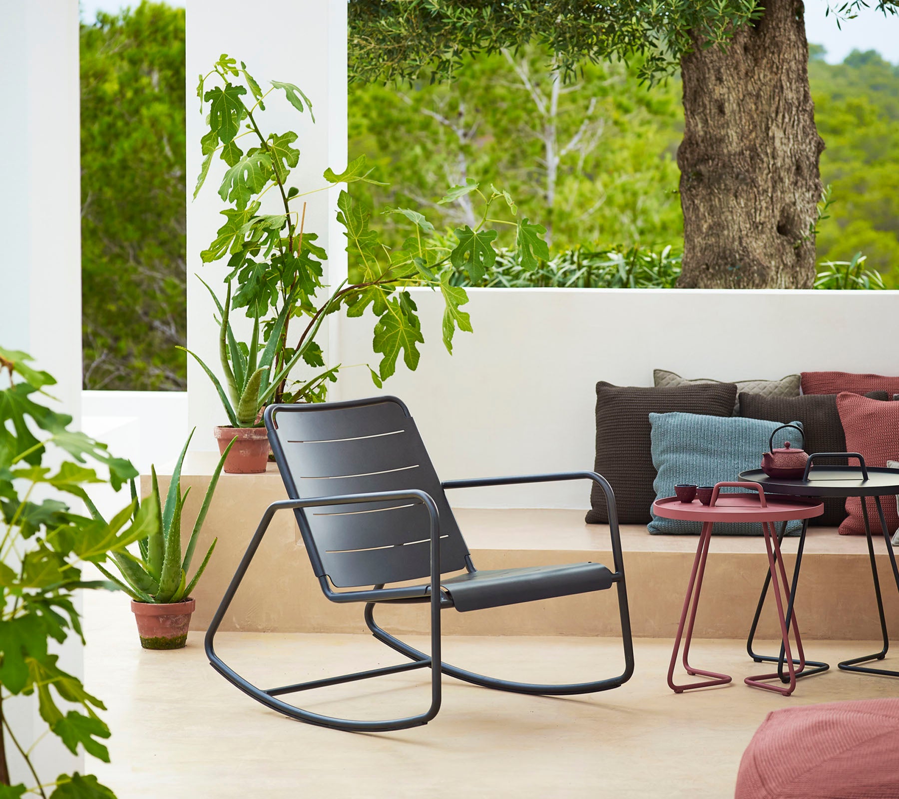 Silla mecedora elegante junto a plantas en macetas y cojines, creando un área de asientos acogedora e invitante con una vista serena.