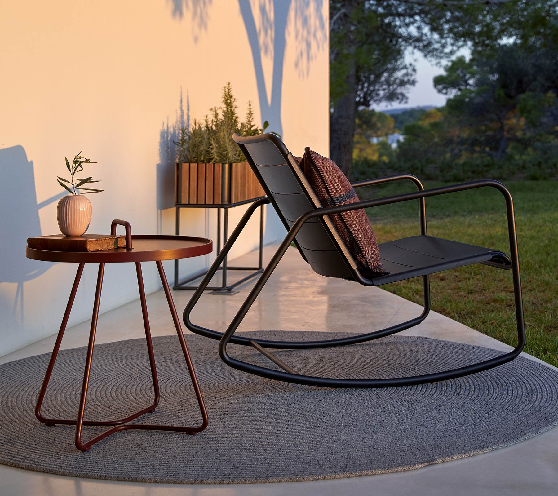 Una silla mecedora elegante con cojines, acompañada de una mesa redonda pequeña y una planta en maceta, contra un fondo sereno.