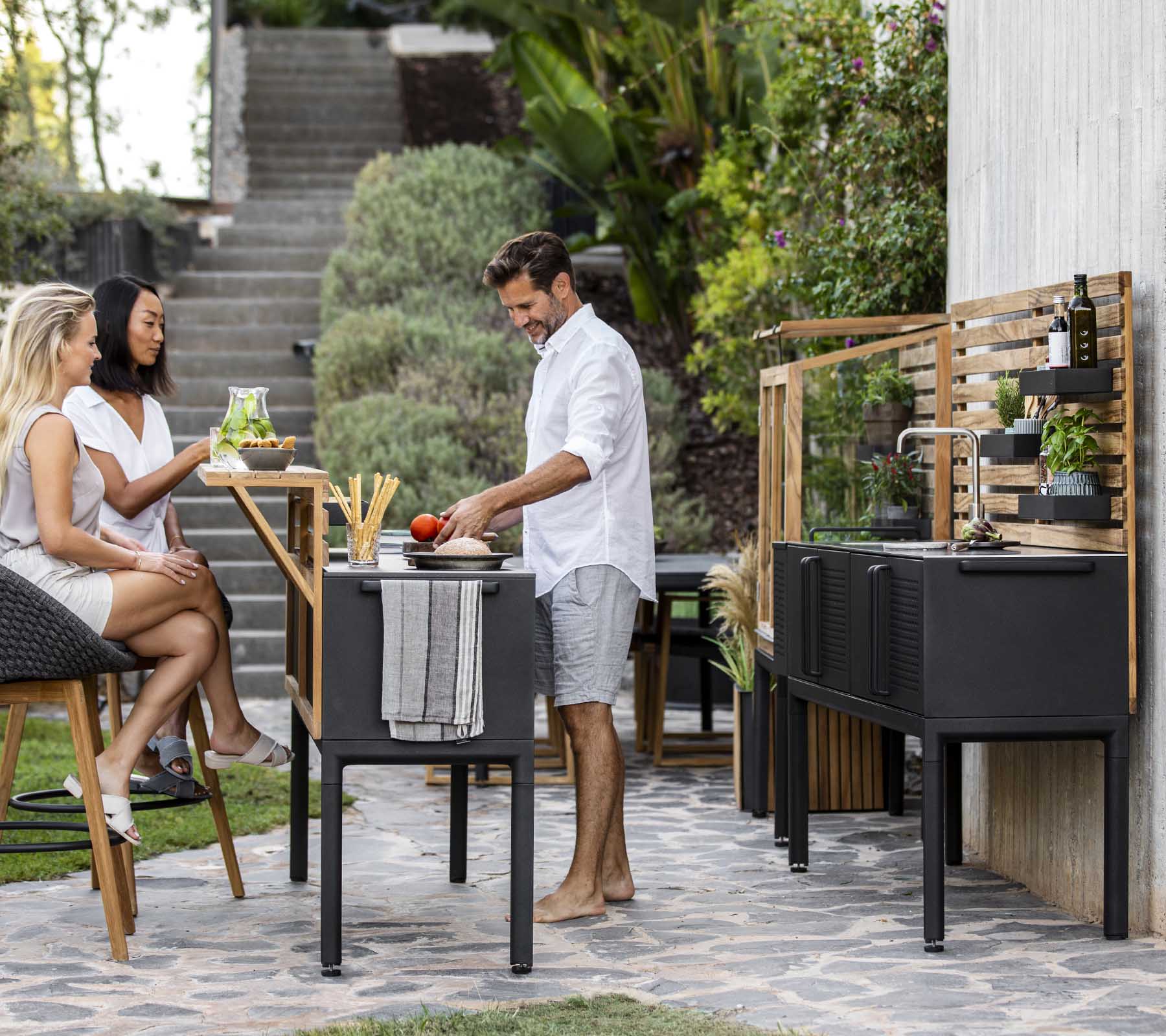 Un hombre prepara comida en una cocina al aire libre moderna, mientras que dos mujeres disfrutan de bebidas cercanas, rodeadas de vegetación y decoración elegante.