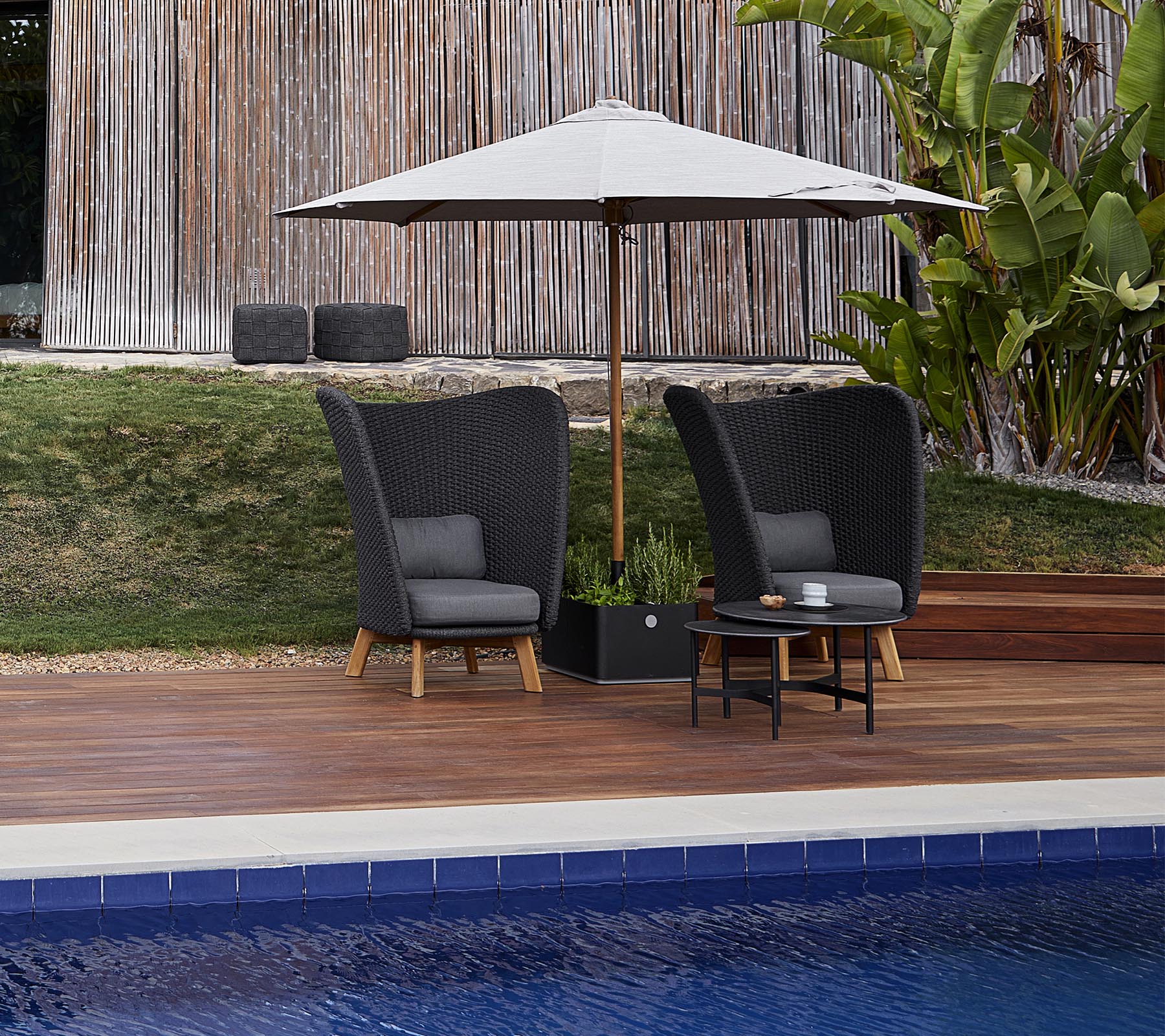 Dos sillas de exterior elegantes con cojines bajo una gran sombrilla, acompañadas de una mesa pequeña, al lado de una piscina.