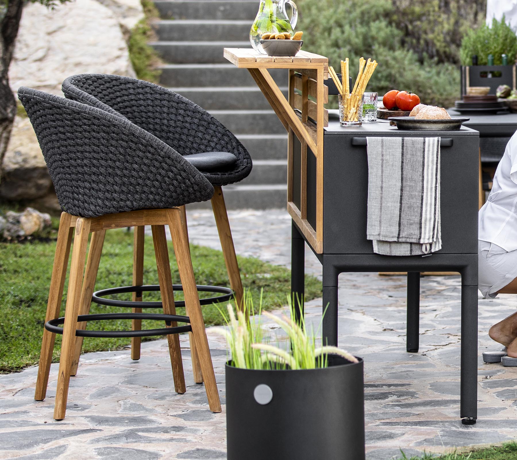 Asientos de exterior elegantes con sillas de mimbre, un carrito de bar moderno y ingredientes frescos dispuestos en una encimera, complementados con plantas decorativas.