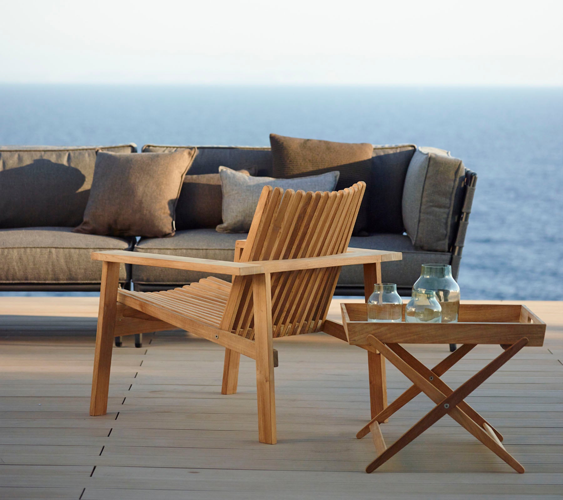 Silla de madera elegante y mesa lateral con botellas, acompañadas de un sofá moderno decorado con cojines, con un fondo de mar tranquilo.