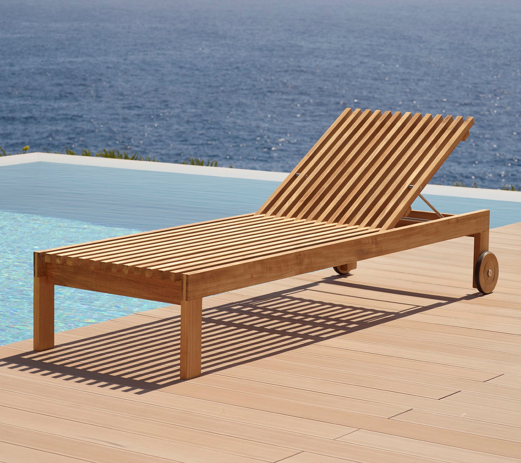 Silla reclinable de madera con diseño de listones, con ruedas y situada al lado de una piscina con vista al agua de fondo.