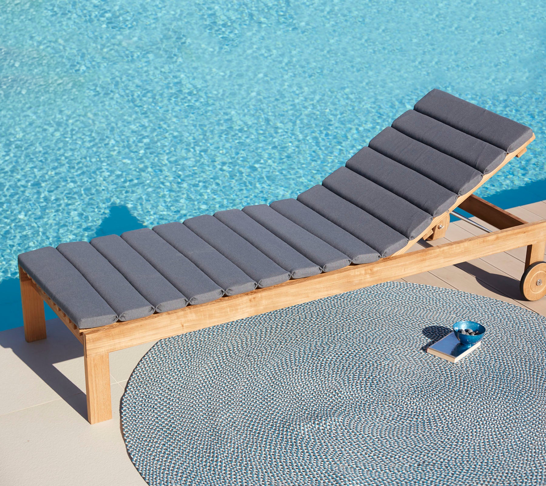 Silla de lounge gris cómoda al lado de una alfombra circular y una pequeña bandeja, con una superficie de agua azul tranquila de fondo.