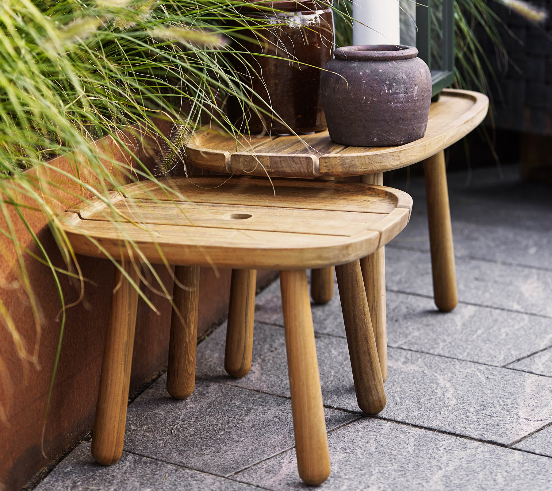 Dos taburetes de madera elegantes en diferentes alturas, complementados por una maceta de cerámica y vegetación en el fondo. Ideal para uso decorativo.
