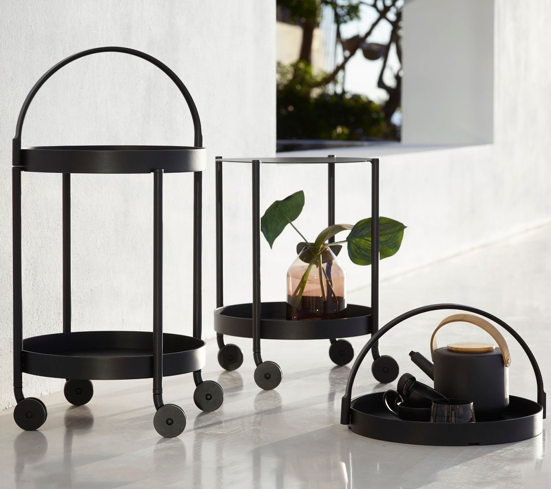 Tres carritos de metal negro con marcos circulares y ruedas, que presentan una planta, elementos decorativos y una taza, organizados para exhibición.