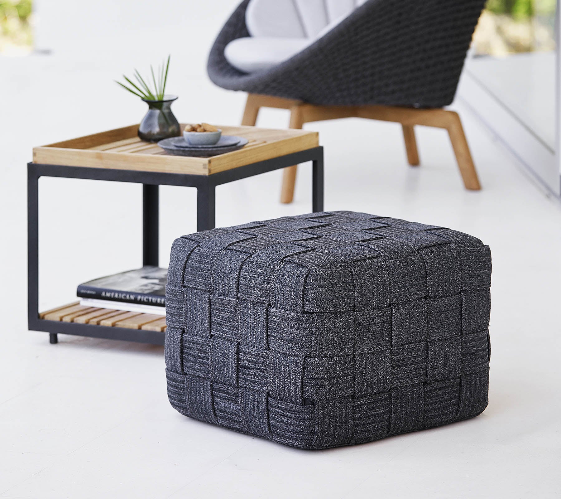 Un pouf tejido gris texturizado situado cerca de una mesa de madera de diseño minimalista, con pequeños artículos decorativos.