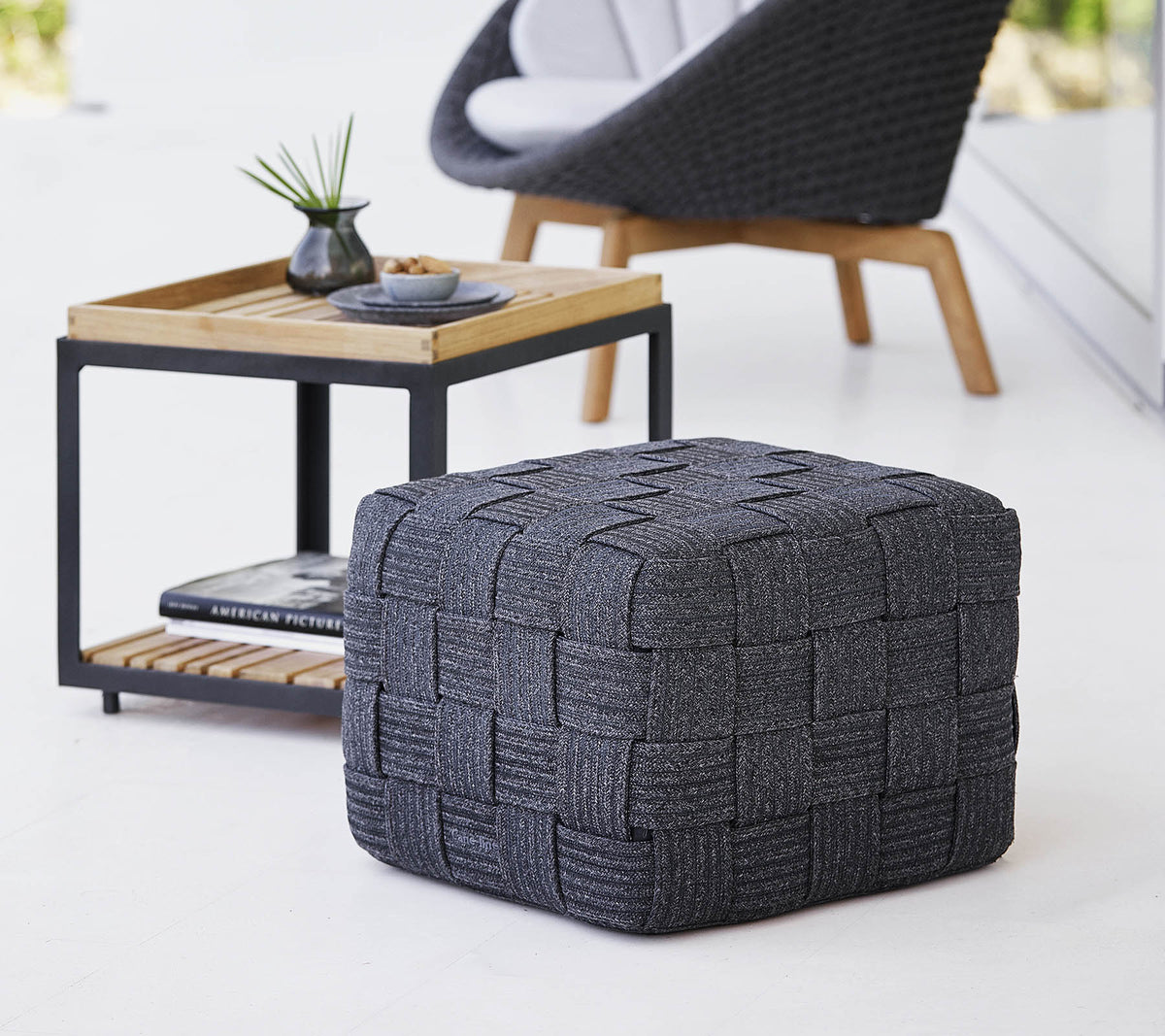Un pouf tejido gris texturizado situado cerca de una mesa de madera de diseño minimalista, con pequeños artículos decorativos.