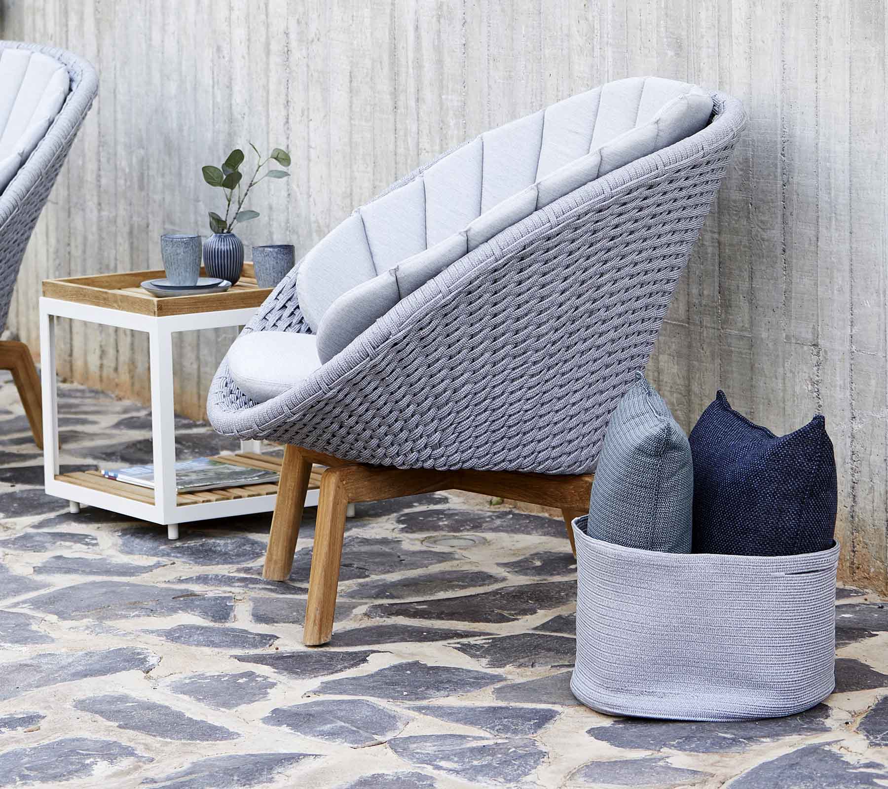 Silla de salón moderna gris con un diseño tejido, acompañada de almohadas y una pequeña mesa lateral de madera. El suelo de piedra mejora la estética.