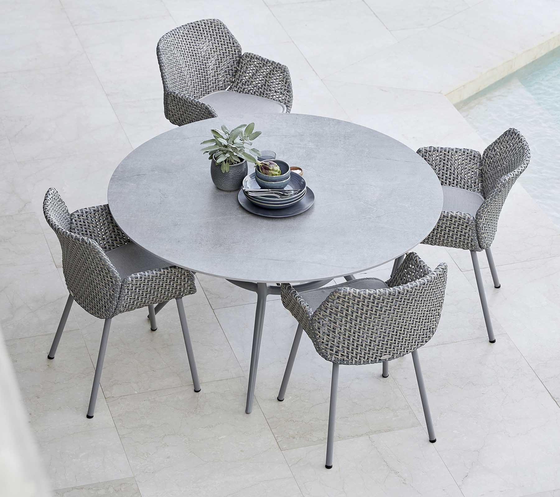 Una mesa redonda elegante combinada con sillas tejidas, con una planta y vajilla en el centro, perfecta para cenar al aire libre.