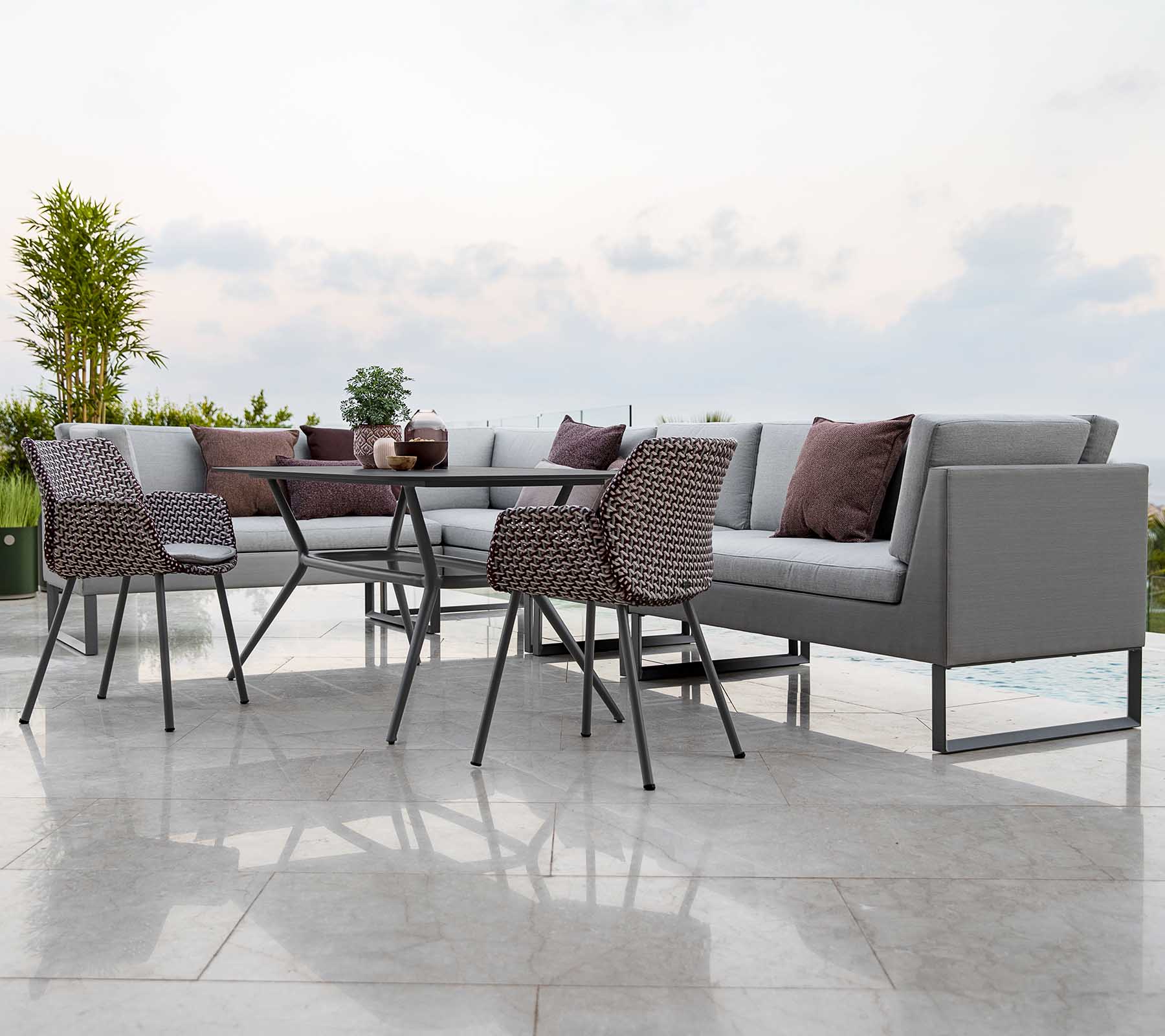 Disposición moderna de asientos al aire libre que incluye un sofá seccional con cojines, una mesa de vidrio y sillas elegantes.