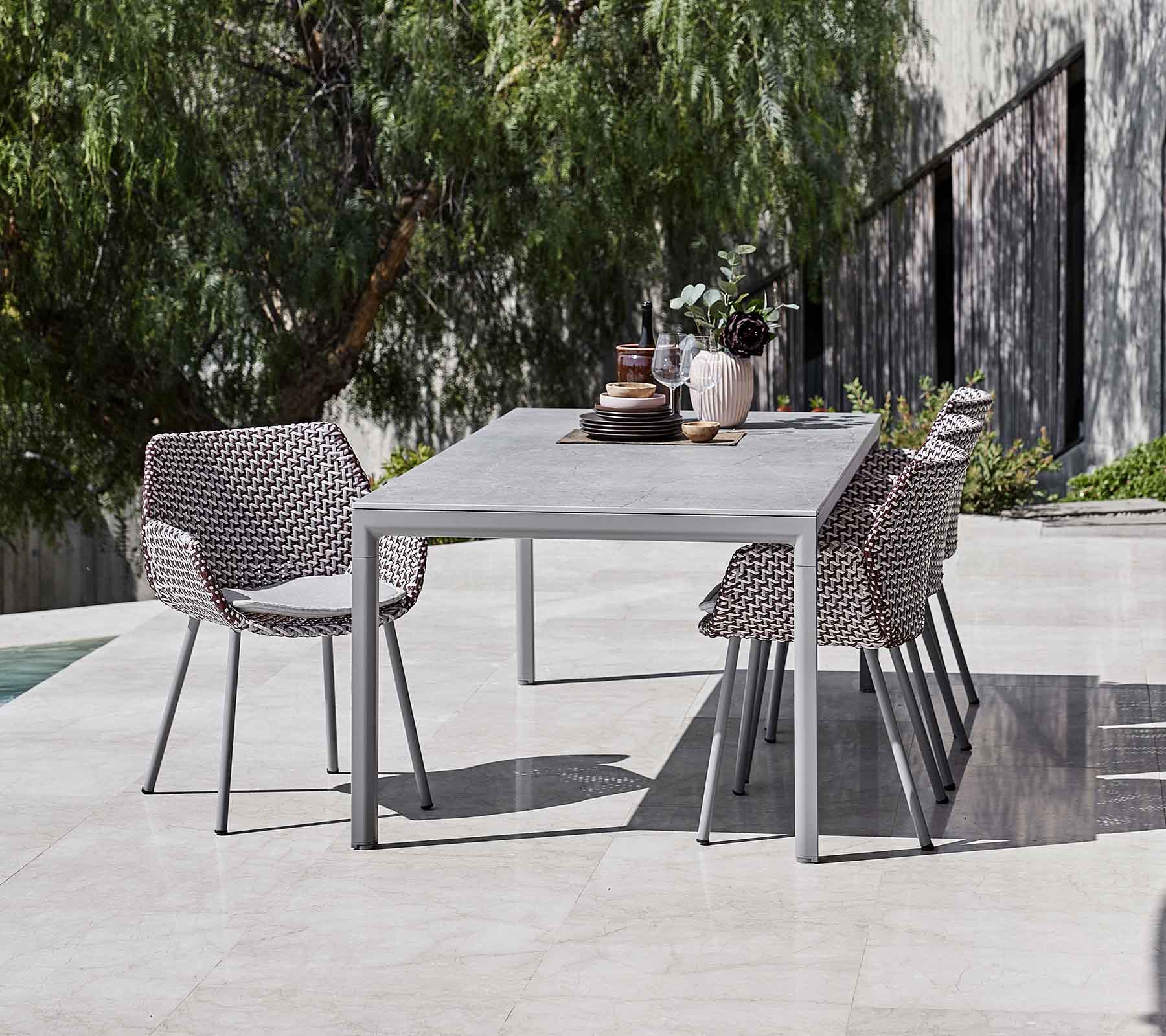 Una mesa de comedor moderna rodeada de sillas tejidas elegantes, complementada con jarrones decorativos y vegetación en el fondo.