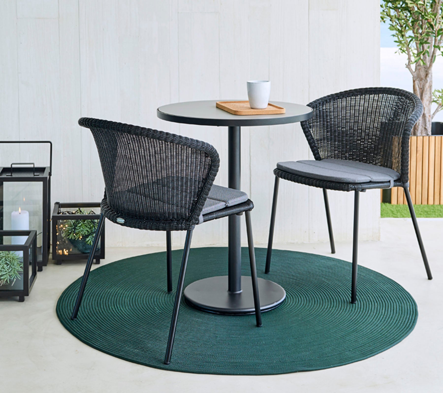 Una mesa redonda con dos sillas de mimbre negras sobre una alfombra verde, acompañada de una taza de café y una tabla de madera. Linternas decorativas en el fondo.
