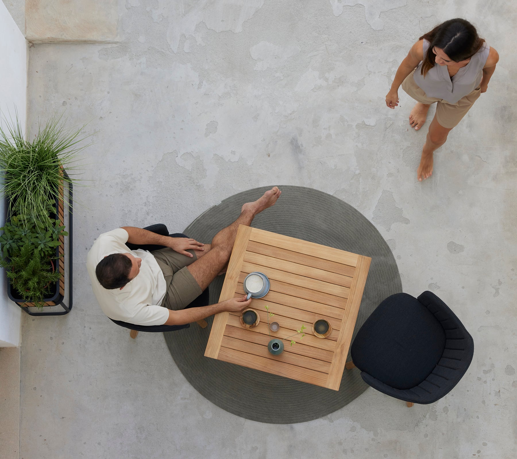 Dos personas disfrutan de café en una mesa de teca rodeada de vegetación, con una silla negra cercana. Se representa una atmósfera relajada de café.