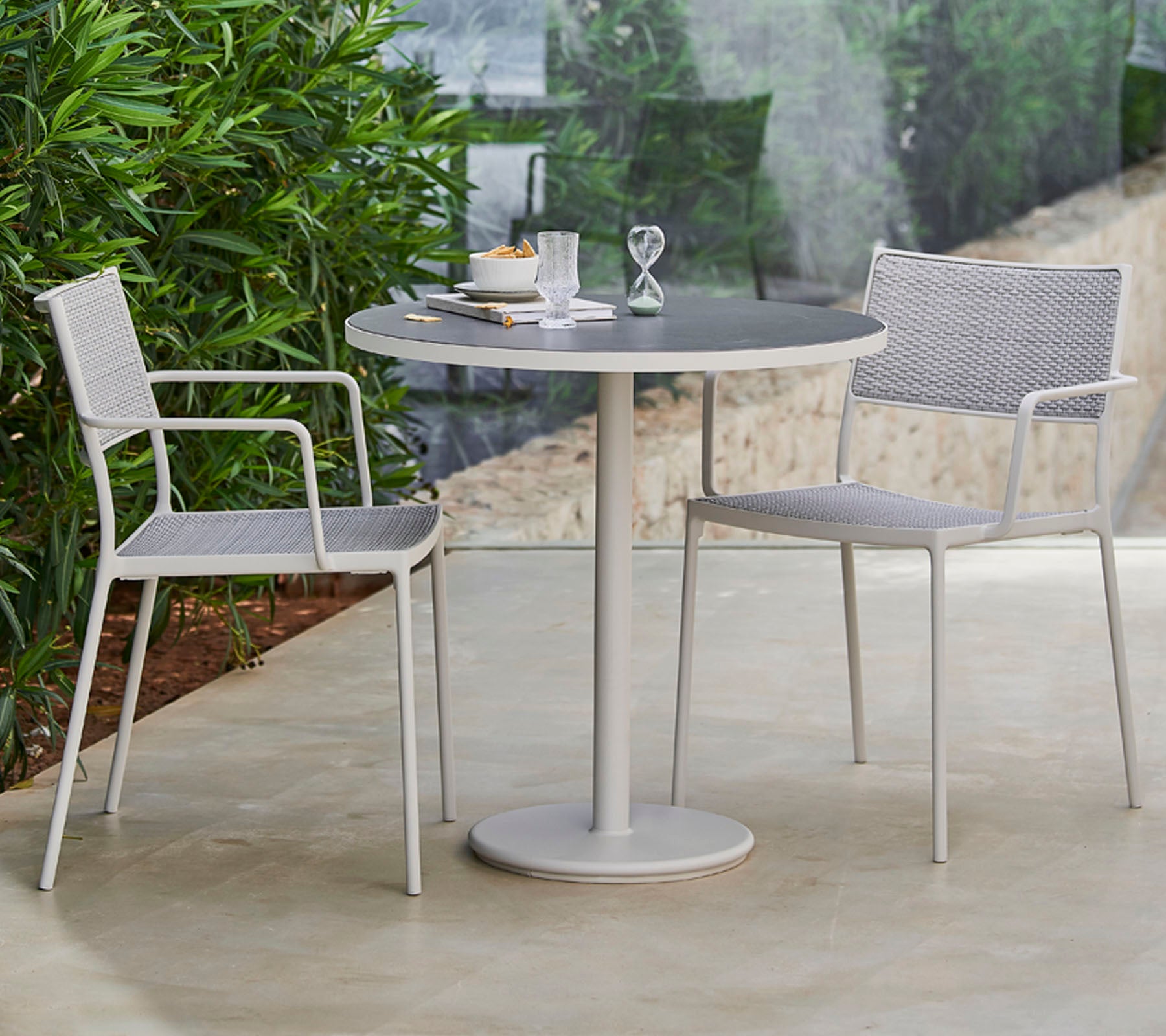 Una mesa redonda elegante rodeada de dos sillas modernas, dispuesta con vajilla y un elemento decorativo, enmarcada por exuberante vegetación.