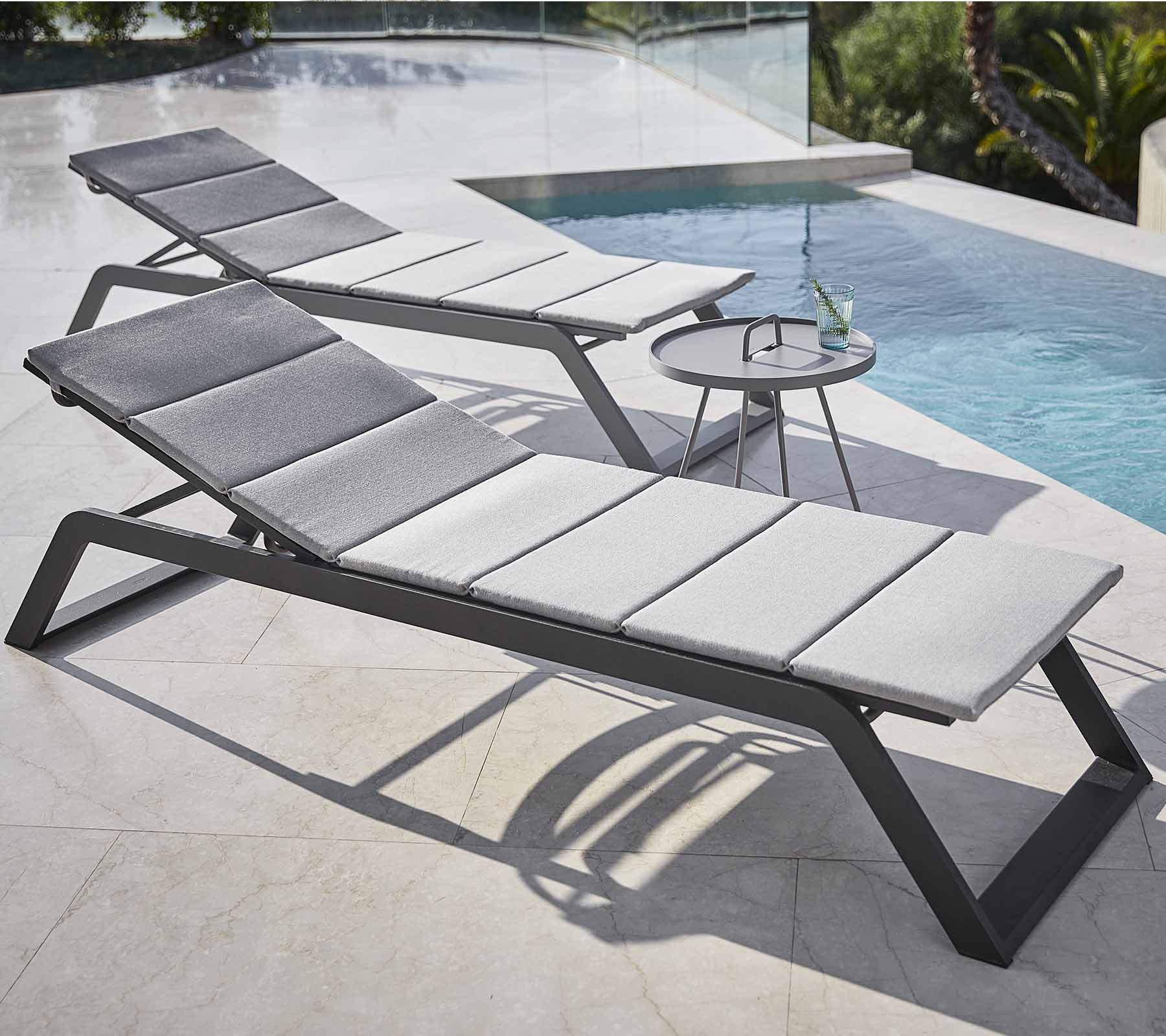 Dos cómodas sillas con cojines grises están situadas junto a una piscina, acompañadas de una pequeña mesa auxiliar con una bebida.