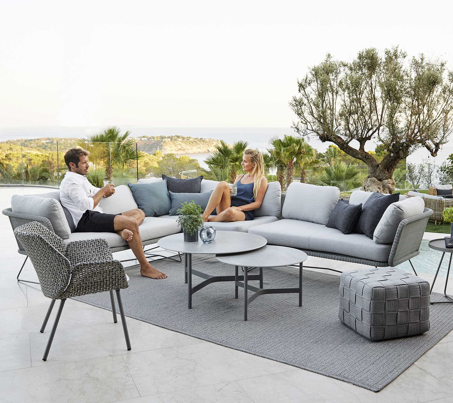 Una moderna área de seating al aire libre con un sofá, un sillón y una mesa de café, donde dos personas se relajan rodeadas de vegetación y una vista escénica.