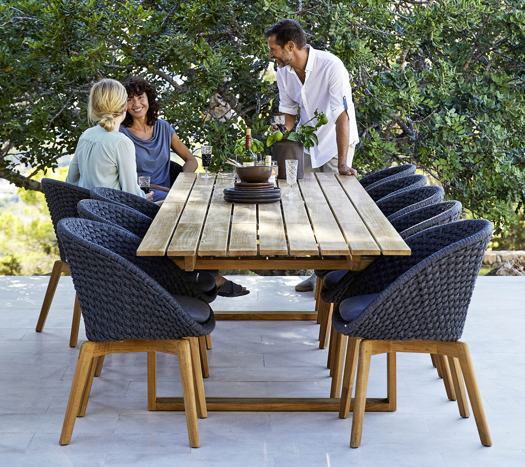 Una elegante configuración de comedor con una mesa de madera rodeada de sillas cómodas, con tres personas participando en una conversación, en medio de la vegetación.