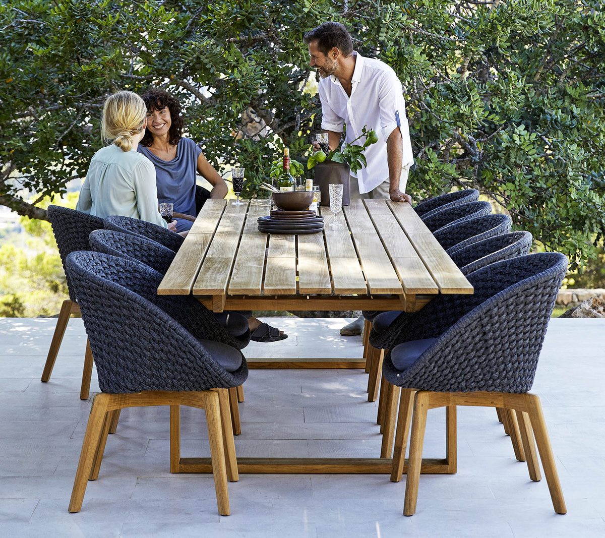 Una elegante configuración de comedor con una mesa de madera rodeada de sillas cómodas, con tres personas participando en una conversación, en medio de la vegetación.