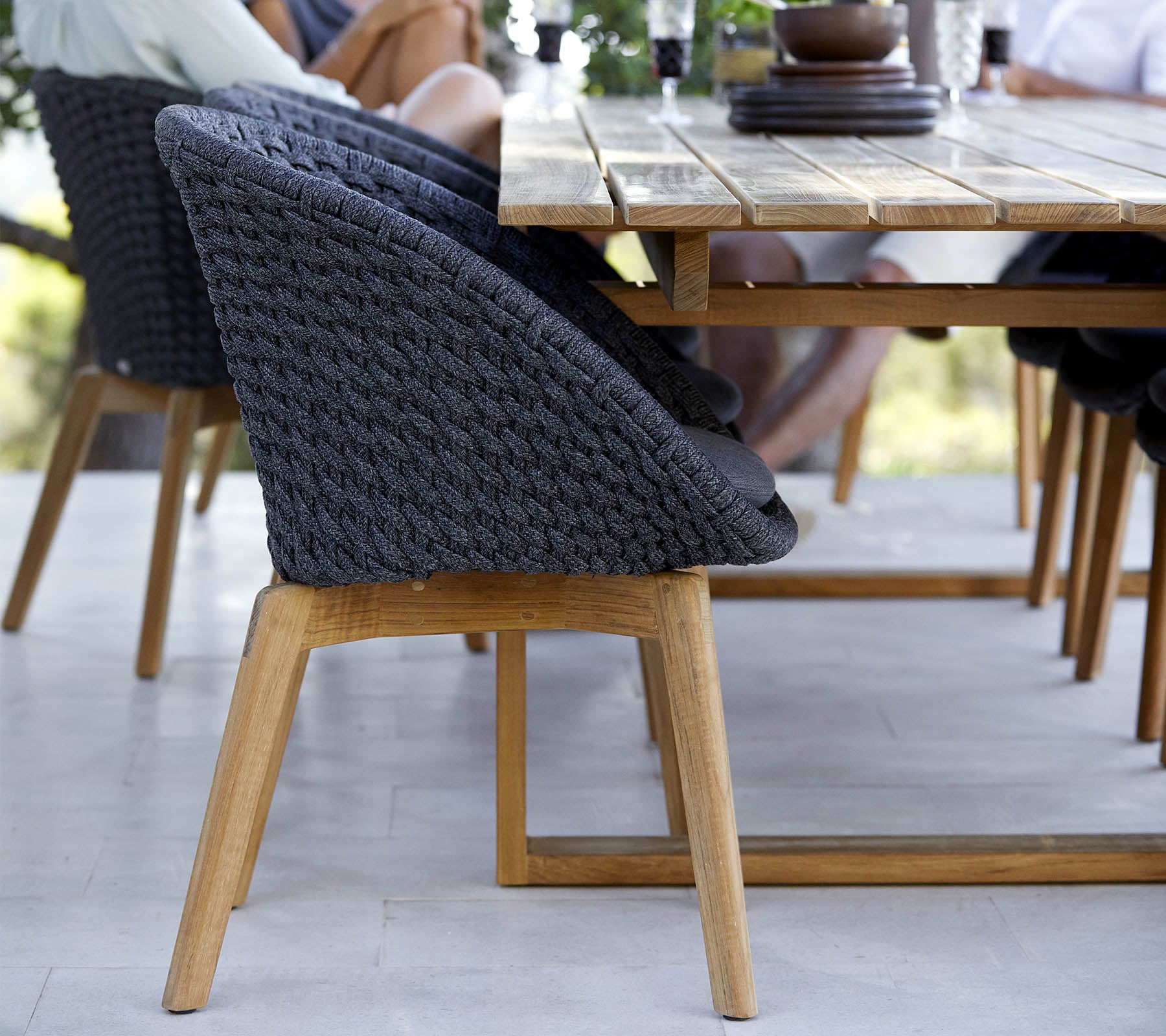 Una silla elegante con respaldo trenzado y patas de madera, situada al lado de una mesa de comedor de madera, con gente comprometida cerca.